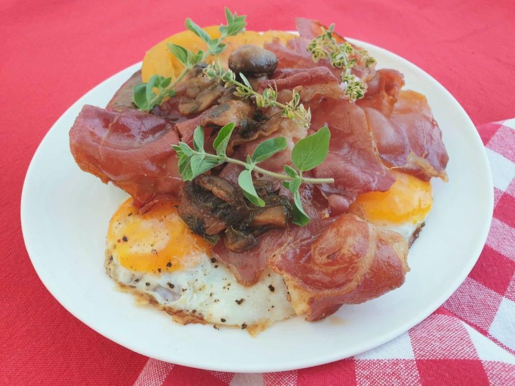 Wit bord vol met gebakken eieren, spek, worstje, tomaten en champignons gegarneerd met verse kruiden